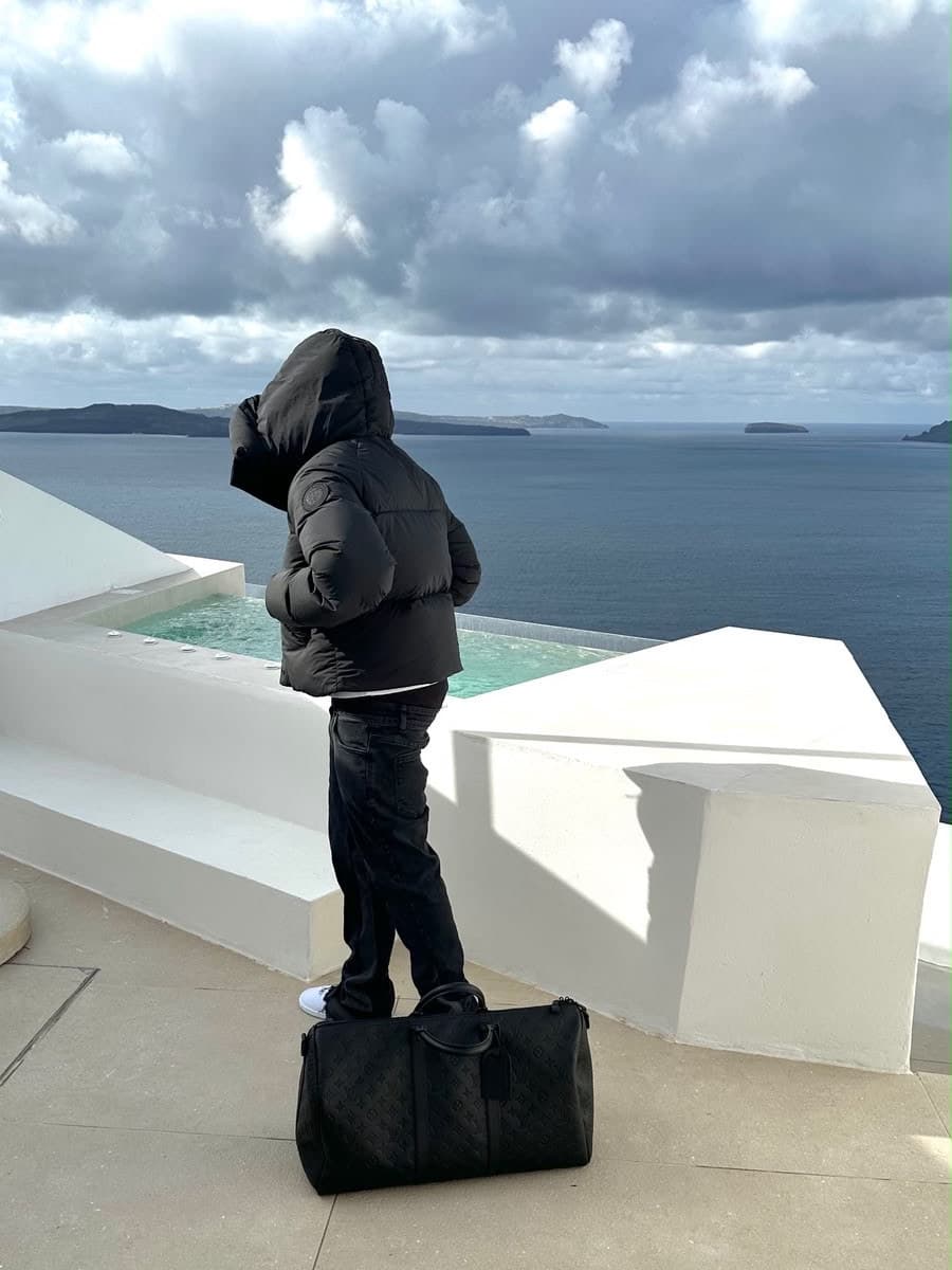 Cris seen from behind on a whitewashed clifftop terrace, in a black hooded puffer jacket and dark jeans, looking out past an infinity pool to the Aegean and the caldera islands under heavy overcast cloud, a black monogrammed duffel bag on the paving at his feet.