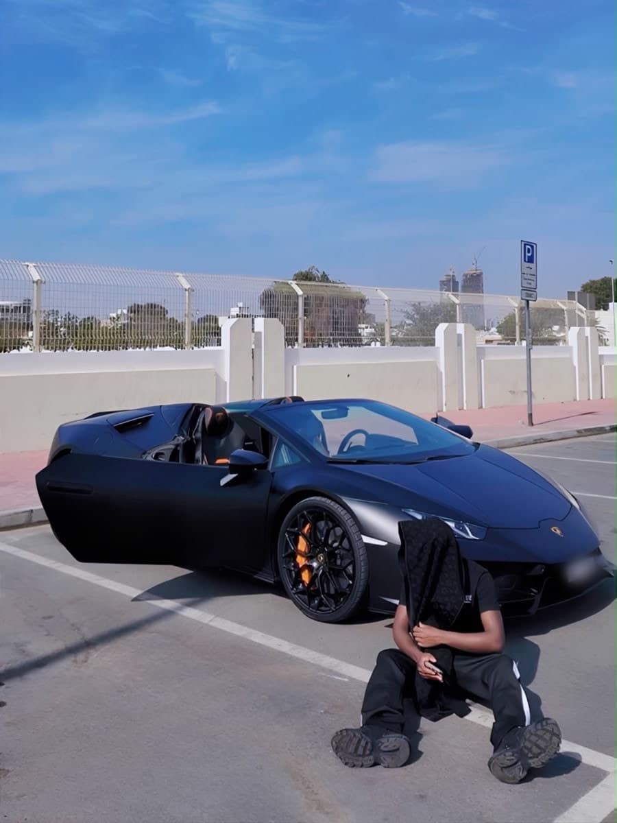 Cris sitting on the asphalt of a marked parking bay in a black tracksuit and chunky trainers, a dark jacket held up over his face, in front of a matte black Lamborghini convertible with its roof down and the driver's door raised; a white perimeter wall, palms and a distant skyline of towers behind.