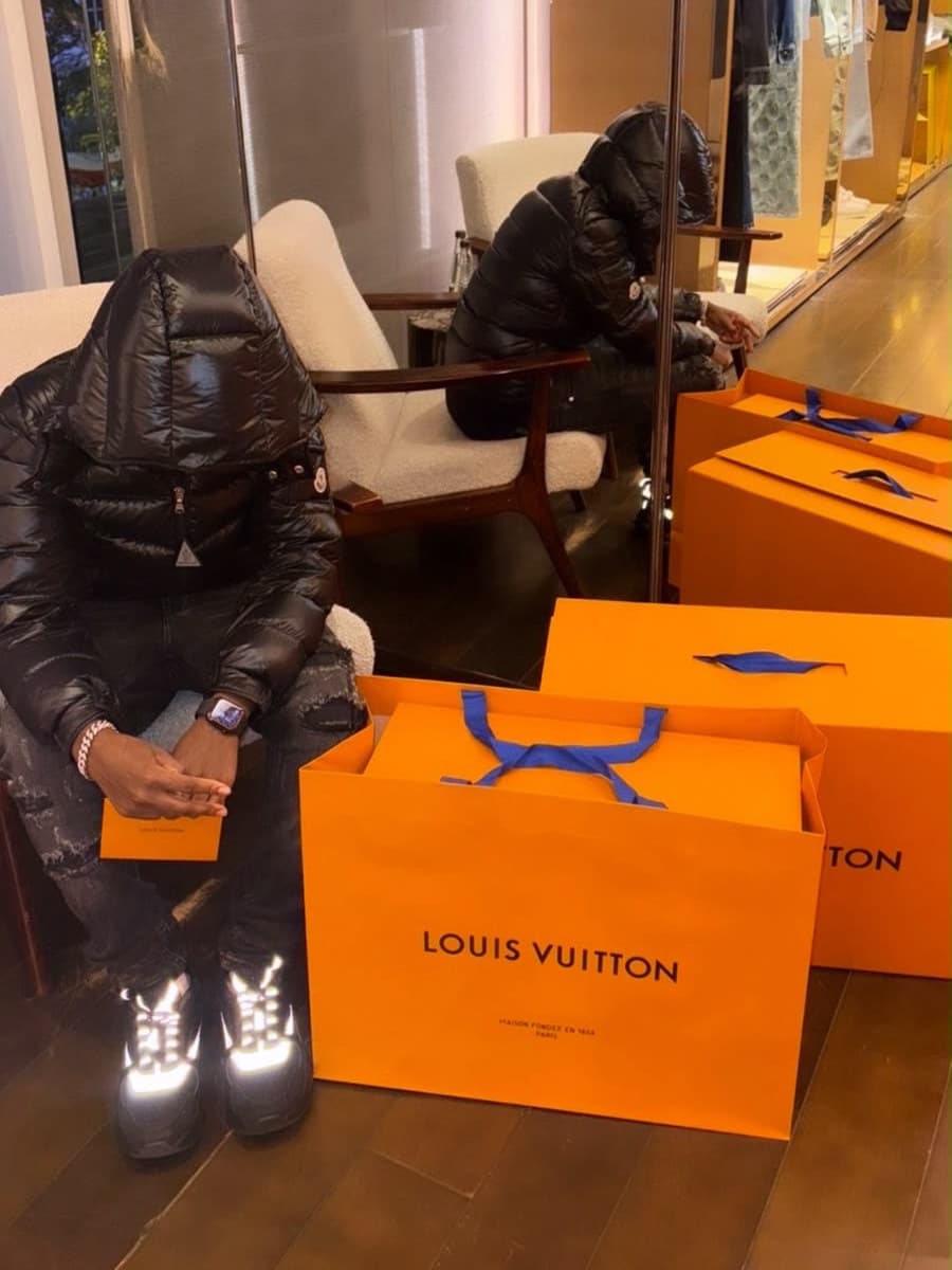 Cris seated in a boutique armchair in a glossy black hooded puffer, the hood pulled down over his face and a small orange card in his hands, a row of large orange Louis Vuitton bags on the dark wood floor beside him and his own reflection in the mirrored partition behind.