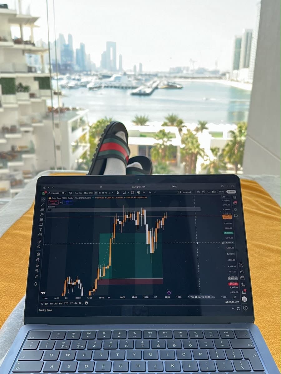 A laptop propped on a yellow towel on a balcony lounger, its screen filled by an hourly XAUUSD candlestick chart (spot gold against the US dollar) with a position tool drawn over the recent range; the photographer's feet in slides beyond the screen, and the Dubai Marina's moored boats, palms and skyline of towers behind.