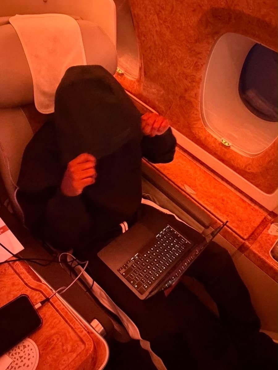 Cris seated in a warmly lit aircraft cabin panelled in burl wood, photographed from above, his face completely covered by a black hood he is holding down with both hands, an open laptop on the seat in front of him, a phone and a white cable on the side table at his left, and an oval cabin window with the blind drawn beyond his shoulder.