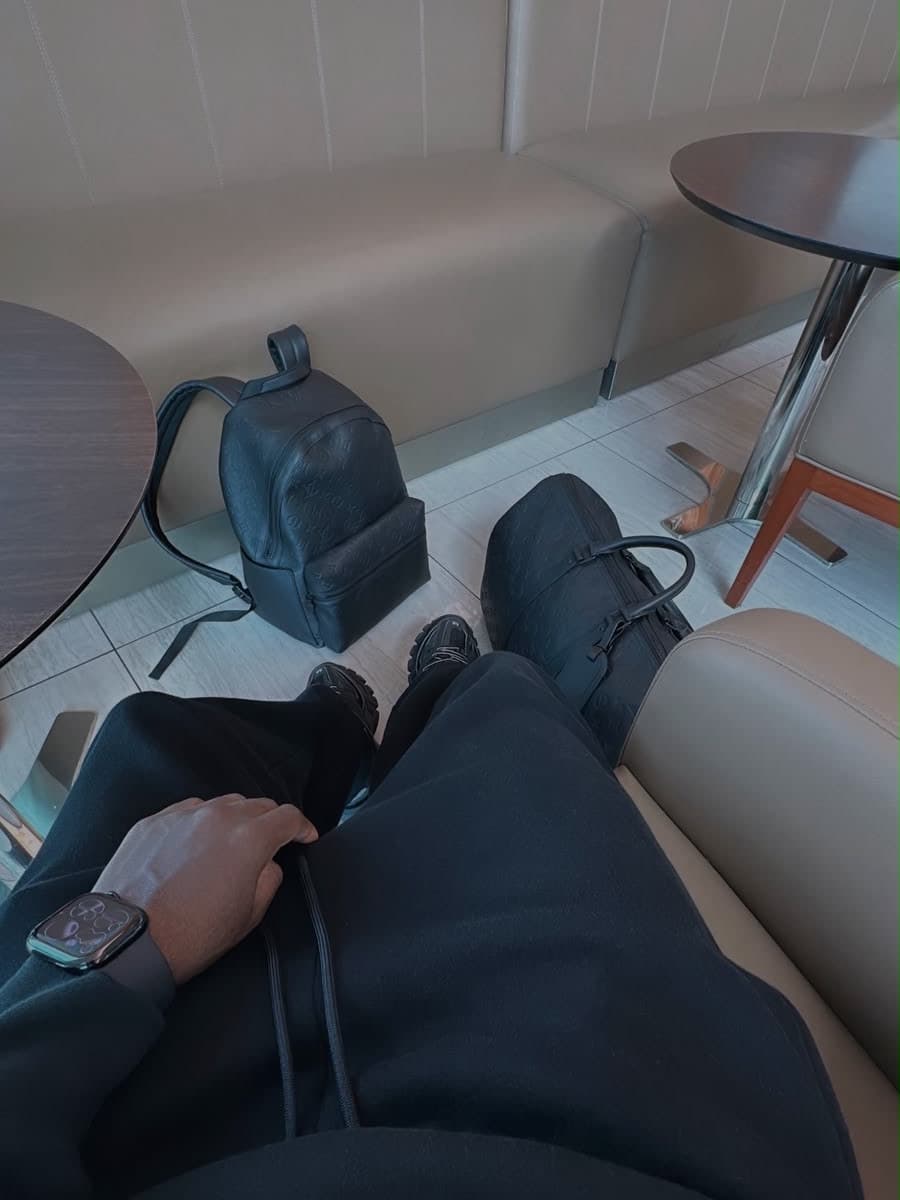 A view looking straight down from an airport lounge seat: black tracksuit, black chunky trainers and a smartwatch on the wrist, with a black backpack in an embossed monogram and a matching holdall on the pale tiled floor between two round pedestal tables and a cream leather banquette.