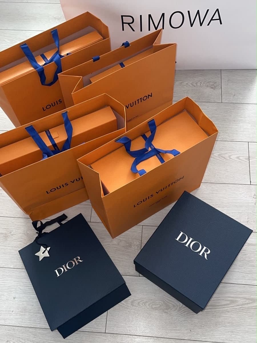 Five orange Louis Vuitton shopping bags with blue ribbon handles, a navy Dior gift bag and a navy Dior box, arranged on a pale wood floor in front of a white Rimowa box.